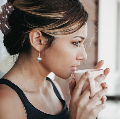 Woman holding a mug, wearing a silver plated puffed heart drop earring on a fishhook wire.