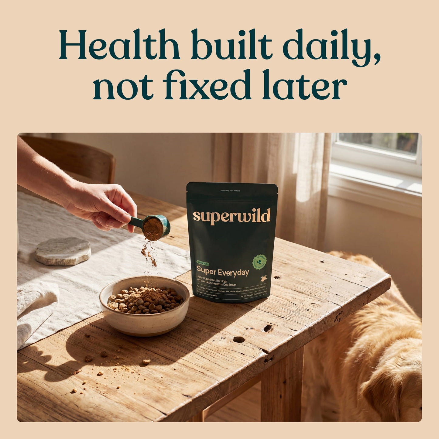 A hand scooping Superwild Super Everyday powder over a bowl of dog food on a wooden table, with a dog resting nearby