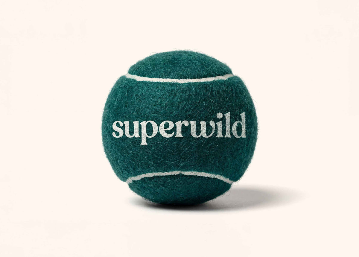 Superwild tennis ball for dogs in deep teal felt, with white curved seam lines and the superwild wordmark printed in white on the side, shown on a plain cream background.